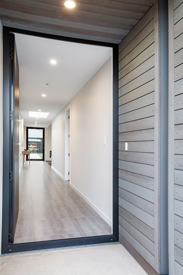 View from an exterior covered entry into a long, bright residential hallway. The entry is framed by grey horizontal timber-style cladding and a dark metal door frame. Inside, the hall has pale tiled flooring, white walls, recessed lighting, a skylight, and glazing at the far end.