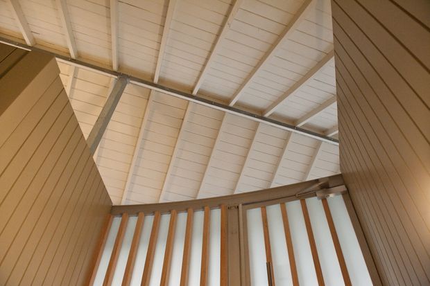 Interior architectural view looking upward toward a high pitched timber-lined ceiling and exposed rafters. A timber balustrade with evenly spaced vertical slats borders an upper-level landing or stair opening, while vertical timber wall panelling frames both sides. The palette is warm natural wood and off-white, with a strong emphasis on timber structure and detailing.