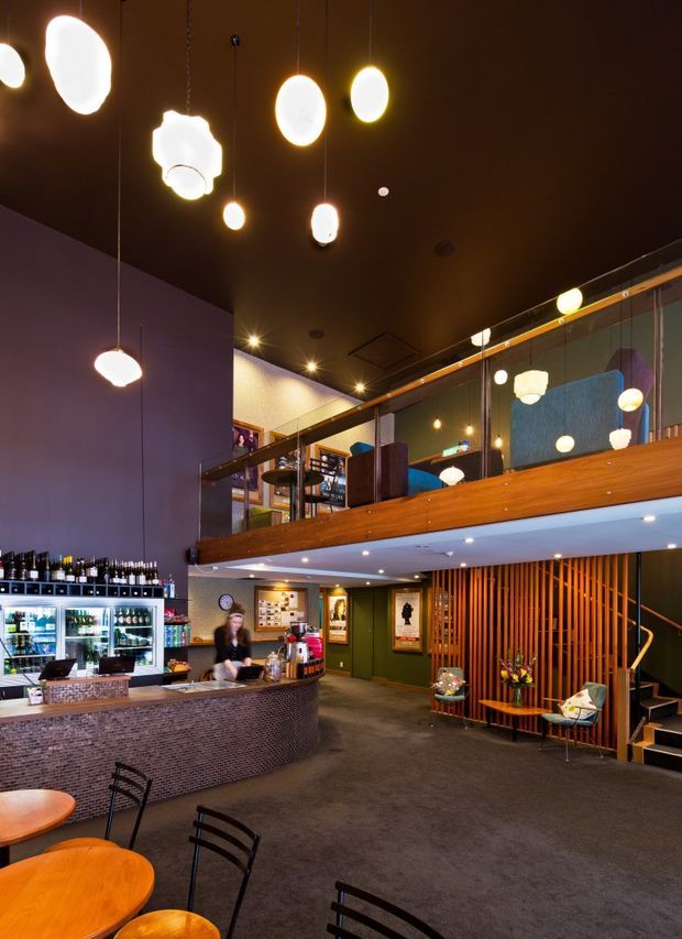 A warmly lit hospitality interior, likely a café, bar, or restaurant lobby. The space features a curved dark tiled service counter with refrigerated drink displays, numerous sculptural pendant lights suspended from a dark ceiling, a high mezzanine with timber-clad balustrade and glass partitions, and a timber slat feature wall beside a staircase. Dark carpet, deep wall colours, timber accents, and intimate seating create a dramatic contemporary atmosphere.