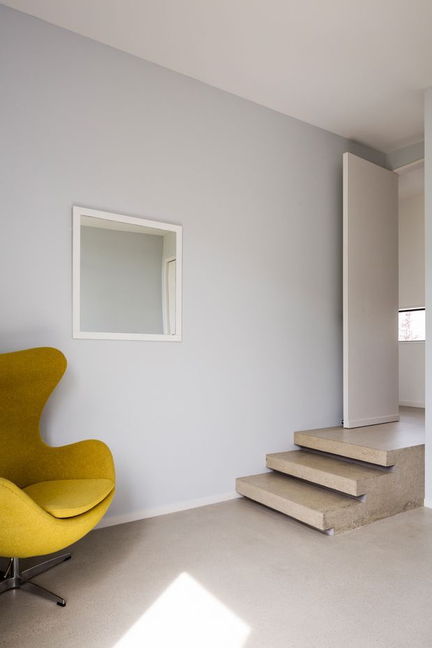 A minimalist interior corner featuring a mustard-yellow sculptural lounge chair, pale grey walls, a small square wall mirror, and a short set of floating concrete or stone-finished steps leading to an upper level. The space is softly lit by daylight, with a restrained neutral palette and a strong geometric composition.