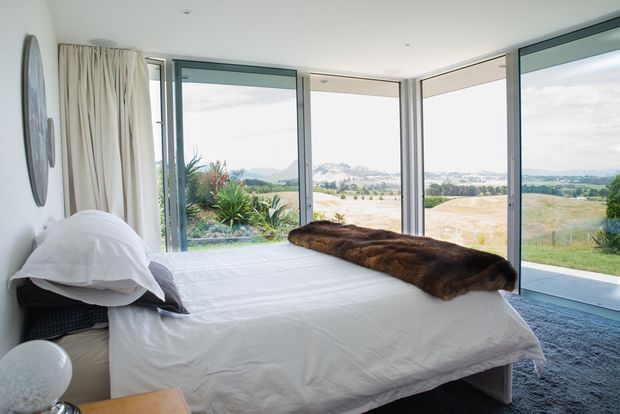 A bright contemporary bedroom with a low-profile bed dressed in white linens and a dark brown fur throw. Full-height corner glazing and sliding glass doors open the room to expansive rural views, while soft curtains, pale walls, and a grey carpet create a calm, understated interior.