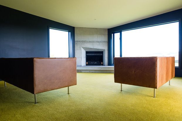 A minimalist living room with two low-profile brown leather armchairs facing a built-in fireplace. The room has a pale green carpet, dark charcoal walls, exposed textured concrete around the fireplace, and expansive dark-framed corner windows providing abundant natural light.