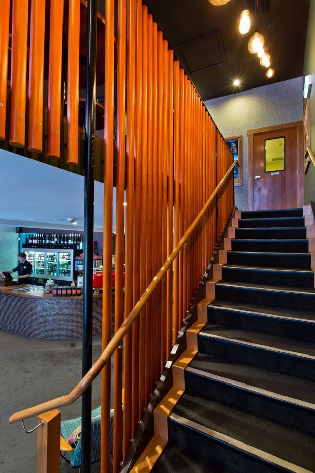 A commercial interior stairway with dark carpeted steps, a warm timber handrail, and a striking screen of closely spaced vertical orange-toned timber slats. The stair rises beside a bar or hospitality area visible below, with a dark ceiling, exposed pendant bulbs, patterned wall surfaces, and a timber door at the upper landing. The image has a warm, dramatic, contemporary hospitality aesthetic.