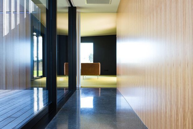 A refined contemporary interior corridor with dark polished stone flooring, a full-height timber-lined wall, and black-framed glass openings on the left overlooking an exterior deck. The corridor leads to a minimalist sitting area with a caramel upholstered bench, dark feature wall, and bright window beyond. Strong linear geometry, reflective surfaces, and natural light create a calm, gallery-like atmosphere.