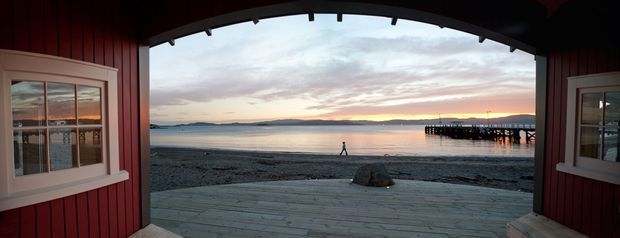 A view from inside a red-painted waterfront boathouse or pavilion through a large open arched opening, looking across a timber deck and pebbly shoreline toward calm water, distant hills, and a pier at sunset. White-trimmed multi-pane windows flank the opening, and exposed dark roof framing is visible overhead.