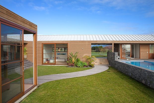 A single-storey contemporary courtyard residence arranged around a landscaped lawn and swimming pool. The building has horizontal timber-clad walls, extensive timber-framed glazing, and a low-pitched light metal roof. A curved paved walkway crosses the lawn toward a covered outdoor area and pool, with a dark rough stone retaining wall forming the pool edge. The view also shows a rural landscape beyond the courtyard.
