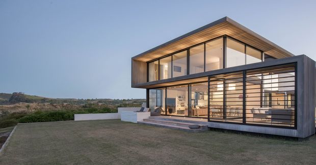 Modern two-storey residence with a rectilinear, cantilevered form, extensive floor-to-ceiling glazing, dark metal framing, and warm timber soffits. The house opens onto a broad lawn and landscaped coastal-looking site, with a covered ground-floor terrace or outdoor living zone visible behind screened glazing. The image is an exterior architectural photograph taken at dusk.