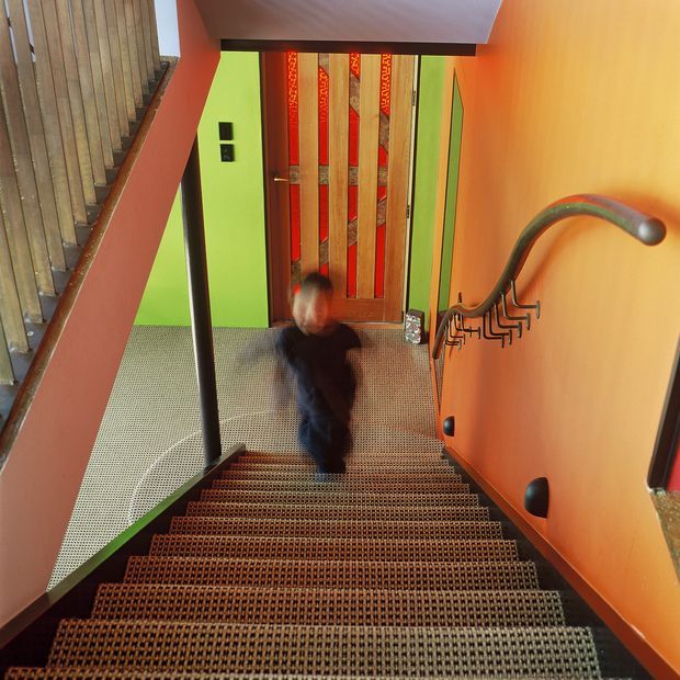 A vibrant interior stairway descending to a landing and timber entrance door. The stairs have dark patterned carpet with metal or dark timber edging, a black handrail and sculptural metal balusters. Walls are painted in saturated orange, green and red tones, with timber wall cladding and a decorative timber-and-glass door. A blurred person is moving on the stairs.