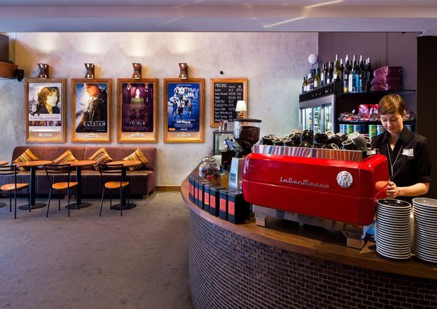 A stylish café or bar interior with a curved service counter in the foreground, faced with small dark mosaic tiles and topped with timber. A bright red commercial espresso machine, stacked cups, wine bottles, refrigerators, and a staff member occupy the counter. The background features booth seating with patterned cushions, small round tables, black chairs, framed posters, and a textured plaster feature wall. The space has warm ambient lighting and a lively hospitality atmosphere.