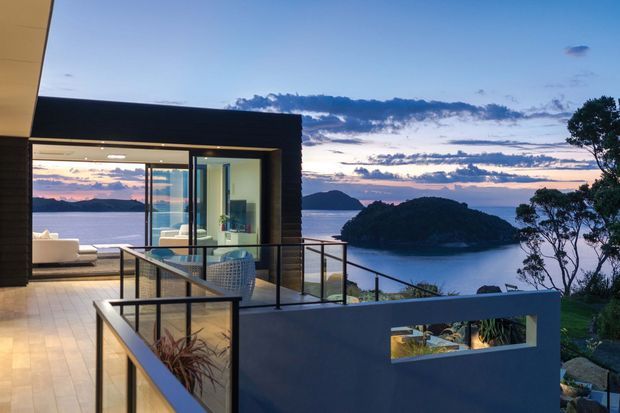 Contemporary coastal residence viewed from an elevated terrace at dusk, with dark horizontal cladding, expansive floor-to-ceiling glazing, and a glass-and-metal balustrade. The open living area is visible through the glazing, while a sheltered bay, wooded islands, and sunset horizon form the background.