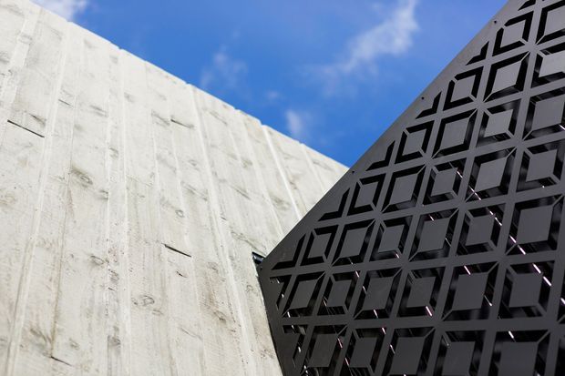 Close-up exterior architectural detail showing a textured white concrete wall intersecting with a dark perforated geometric screen or facade panel. The patterned screen features repeating angular polygonal openings, set against a bright blue sky, creating a strong contrast between raw concrete and black metalwork.