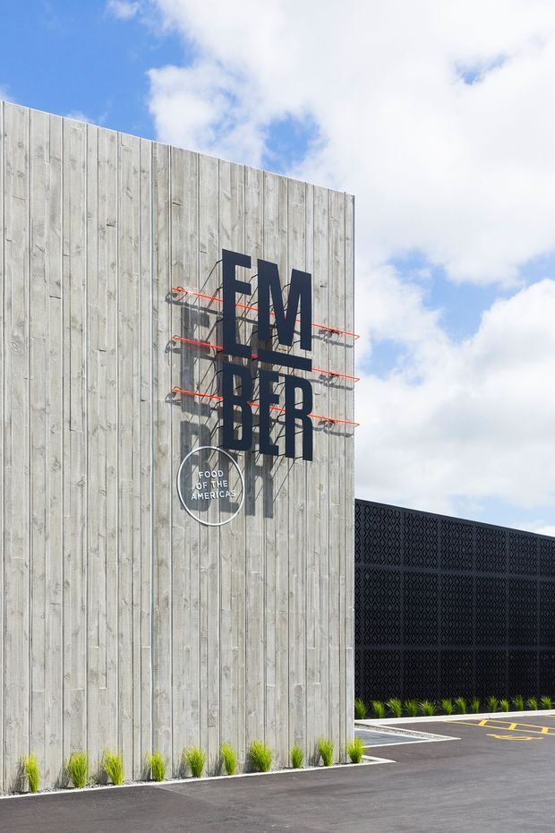 Exterior view of a contemporary commercial building featuring a tall vertical weathered timber-clad facade with large projecting black “EMBER” signage and a circular “Food of the Americas” logo. A contrasting black patterned screen wall extends to the right, with landscaped grasses, paved parking, and a bright partly cloudy sky.