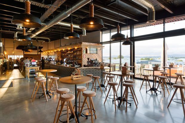 A spacious industrial-style café or bar interior with polished concrete flooring, exposed dark ceiling structure, visible ductwork, hanging pendant lights, and large floor-to-ceiling windows. The room contains numerous timber-topped tables, stools, and high communal tables, with a fully fitted bar and display shelving at the rear. Bright daylight and a waterfront or harbour landscape are visible through the glazing.
