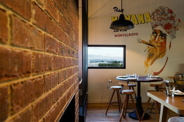 A warmly lit restaurant or café dining area with exposed red brick walls, timber flooring, timber tables and stools, and black metal pendant lighting. A large illustrated mural dominates the wall, while a window provides a view of the landscape outside.