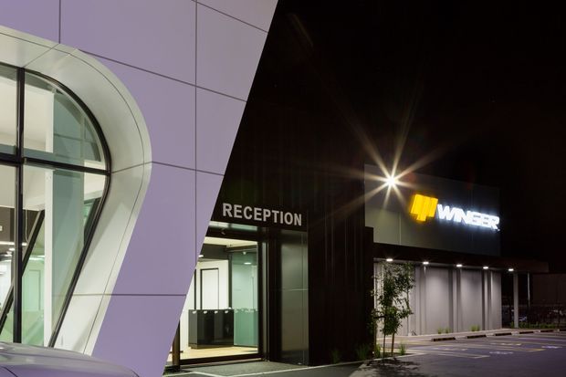 Night-time exterior view of a contemporary commercial building, showing a prominent curved glazed entrance facade on the left, a recessed glass reception entry, and a dark-clad illuminated showroom or retail frontage with signage on the right. The paved forecourt and exterior lighting are visible.
