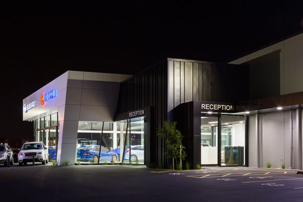 Night-time exterior photograph of a contemporary automotive dealership or commercial showroom. The building has illuminated reception entrances, extensive floor-to-ceiling glazing displaying vehicles, angular light-coloured cladding, dark vertical facade panels, and a paved parking area with marked accessible spaces.