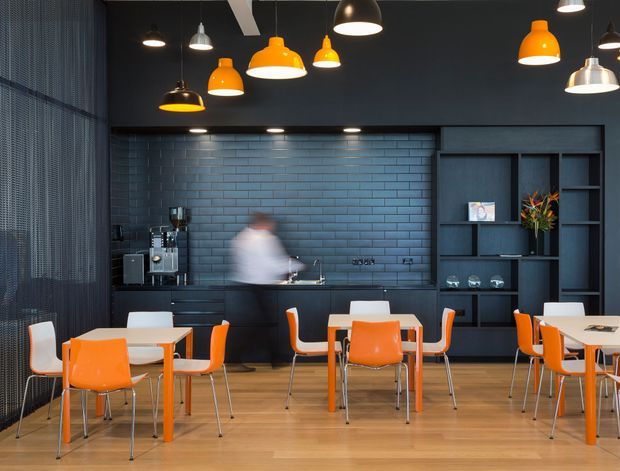 A stylish contemporary commercial café or dining space with a dark blue-black interior, exposed blue subway-tile backsplash, extensive built-in shelving and counter joinery, and a series of colourful pendant lights. White tables and bright orange chairs are arranged across a warm timber floor, with a service counter and coffee equipment along the rear wall.
