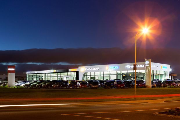 Night-time exterior photograph of a large contemporary automotive dealership or commercial showroom. The long, brightly illuminated glazed facade displays multiple vehicle-brand signs, with a tall roadside pylon sign, extensive car parking, street lighting, and light trails from passing traffic in the foreground. Dark hills form the distant background.