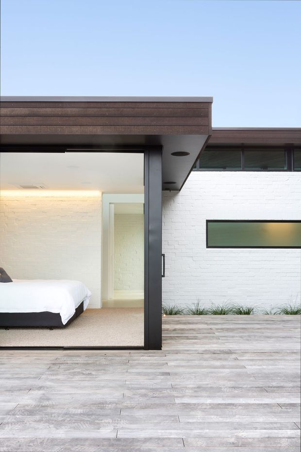 Contemporary courtyard-facing bedroom with white-painted brick walls, a low-profile dark-framed glass opening, and a broad weathered timber deck. The partially visible bedroom has a white bed, pale carpet, and warm concealed lighting, while projecting timber-clad roof forms and dark metal framing create a clean modern architectural composition.