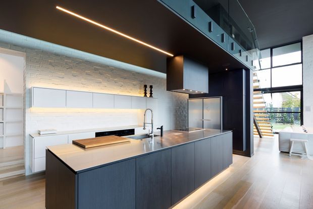 Contemporary open-plan kitchen with a large dark timber island, white flat-panel cabinetry, integrated appliances, a timber benchtop section, and a textured white brick splashback. The space features a dramatic dark ceiling with linear LED lighting, exposed structural and service elements, and full-height glazing leading to a bright adjoining area and stairway.