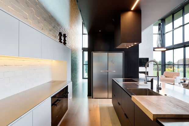 A spacious contemporary kitchen with a long dark island, timber benchtop, integrated appliances, white cabinetry, exposed white-painted brickwork, and full-height black-framed glazing. A large sculptural rangehood and linear lighting contribute to the industrial-modern character, while a living area is visible beyond the kitchen.
