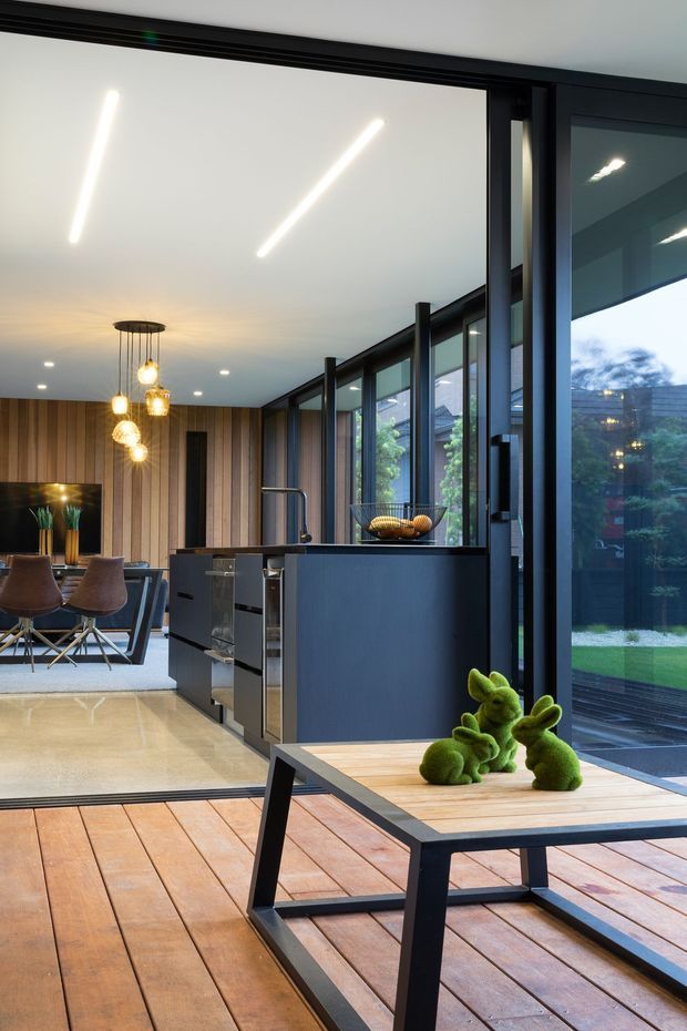 A contemporary open-plan kitchen and dining area opening onto a timber deck through extensive black-framed glazing. The space features a dark kitchen island, vertical timber wall panelling, polished light flooring, modern linear ceiling lights, pendant lighting, and a dining table with sculptural green decor. An outdoor garden is visible through the glass.