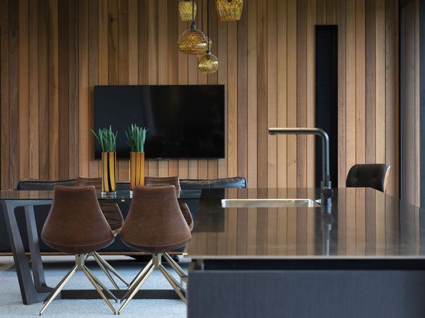 A sophisticated open-plan kitchen and dining area featuring a dark stone island with integrated sink and sculptural tap, a glossy dark dining table, brown leather dining chairs, and a wall-mounted television. The room is defined by extensive vertical timber wall panelling, dark cabinetry and surfaces, and decorative woven pendant lights.