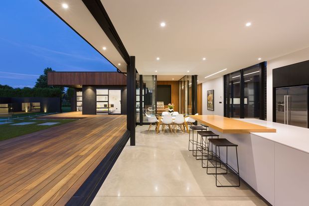 A refined contemporary open-plan kitchen and dining area opening onto a large timber deck and landscaped outdoor space. The interior features a long pale island with integrated seating, white cabinetry, dark-framed full-height glazing, polished light flooring, recessed lighting, and warm timber accents. A separate modern pavilion-like structure is visible outside.