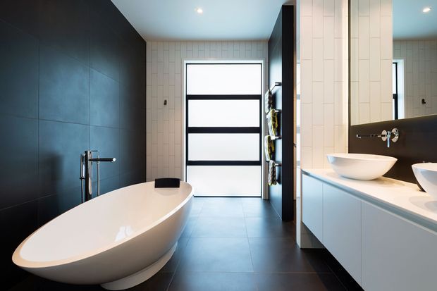 Contemporary bathroom with a freestanding white oval bathtub, twin white vessel basins on a pale floating vanity, dark tiled feature walls, light gridded wall tiles, a large frosted-glass door with black framing, and dark floor tiles. The space has clean lines, recessed lighting, and a restrained monochromatic palette.