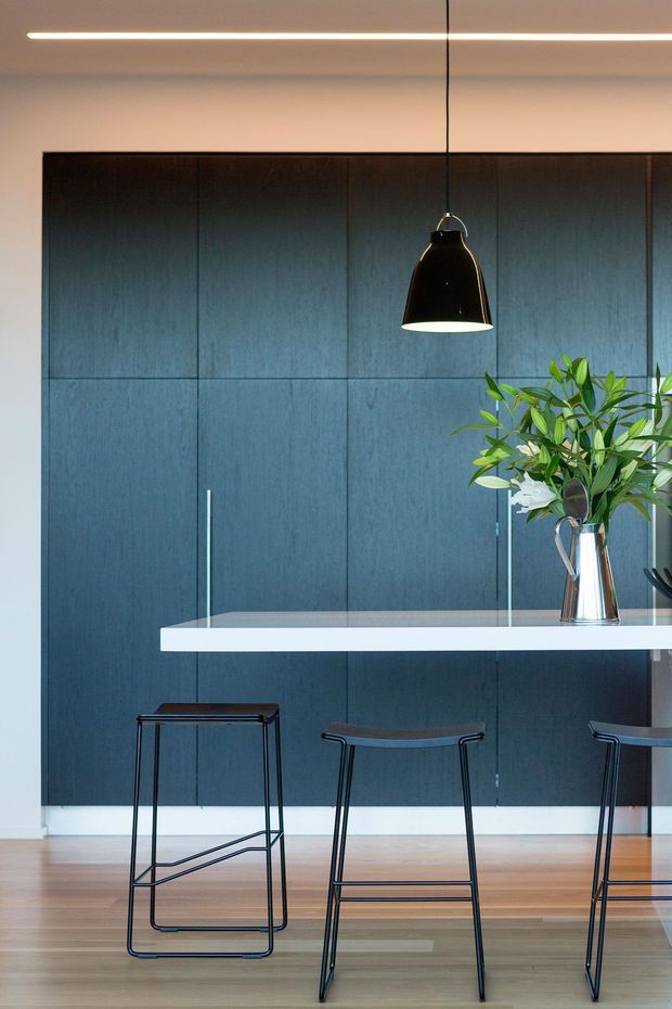 Contemporary interior featuring a long white kitchen island with slim black bar stools, a dramatic dark blue-black timber-panelled wall, warm timber flooring, and a black pendant light. A vase of green foliage adds a natural accent, while linear ceiling lighting provides a clean, refined finish.