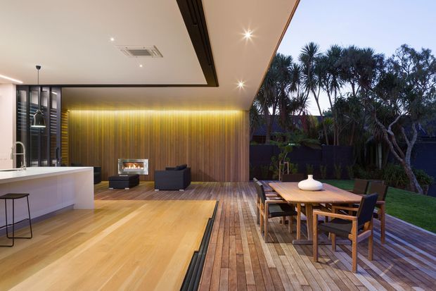 A sophisticated indoor-outdoor living and dining area with large sliding openings connecting a contemporary kitchen and lounge to a timber deck. The interior features warm timber flooring and vertical timber wall panelling, a linear fireplace, recessed lighting, and a minimalist kitchen island. The deck has a timber dining table and chairs overlooking landscaped planting and lawn.