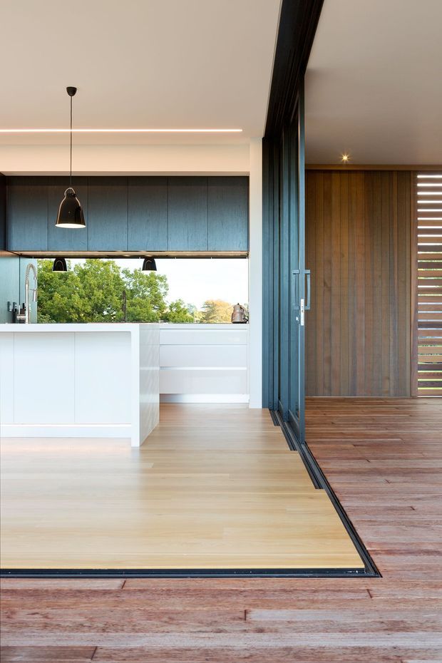 A bright contemporary open-plan kitchen viewed from a timber-lined outdoor deck or terrace. The kitchen features a white island and cabinetry, dark blue-grey overhead cabinetry, pale timber flooring, integrated linear lighting, pendant lights, and a large opening with dark-framed folding or sliding glazing connecting to the deck. The adjoining exterior wall and deck are clad in warm vertical timber, with horizontal louvres at the right.