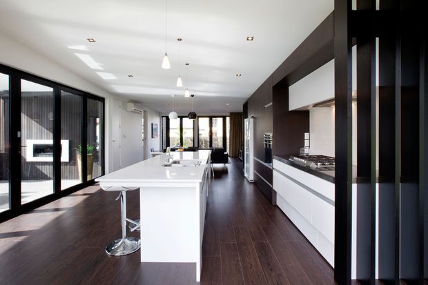 A bright, elongated contemporary kitchen with a large white island and integrated seating, dark timber flooring, extensive black-framed glazing, and sleek white cabinetry. A dark timber feature wall and vertical black screening frame the right side, while pendant lights and recessed ceiling lights illuminate the space.