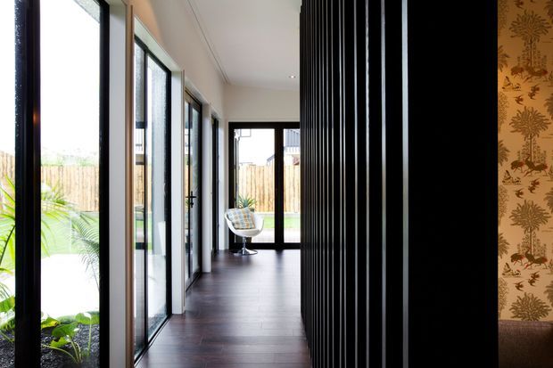 A long, contemporary residential hallway with dark timber flooring, extensive black-framed floor-to-ceiling glazing on one side, and a dramatic vertical black slatted partition on the other. A patterned botanical wallpapered wall and partial upholstered seating appear at the far right, while a bright garden courtyard is visible through the glazing.