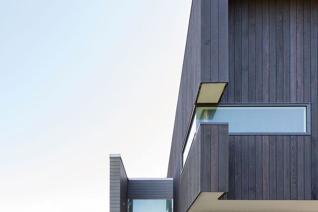 Close-up exterior view of a contemporary multi-level building with dark vertical timber cladding, projecting box forms, deep overhangs, and a narrow horizontal window. The composition emphasizes sharp geometric massing and restrained modern detailing against a clear sky.