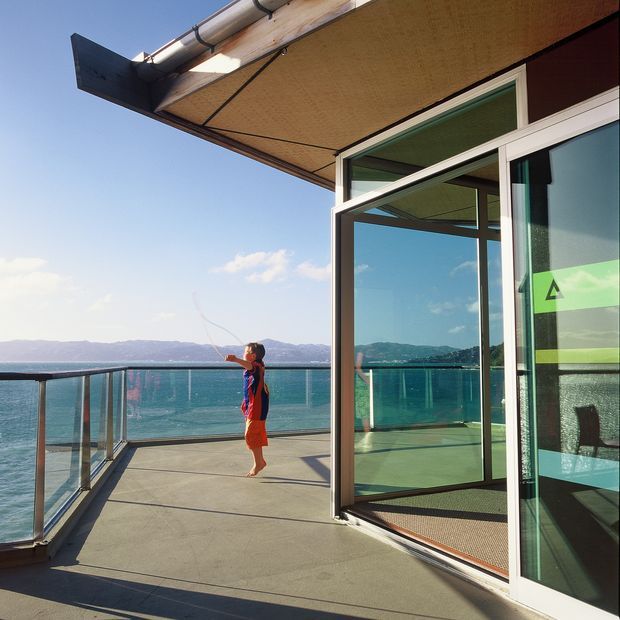 Sunlit modern waterfront terrace or balcony with frameless glass balustrades, concrete paving, timber soffit, exposed gutters, and large white-framed glazed doors opening into the interior. A child stands on the terrace, with turquoise water and distant hills beyond. The image emphasises the outdoor living space and its expansive water views.