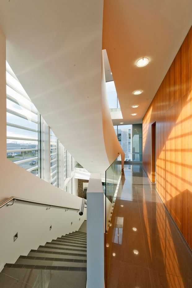 A bright, multi-level interior stairway and landing in a contemporary building. Broad glazed windows line the left side, bringing in daylight and views of an urban setting. The stair has dark grey tiled treads, a metal handrail, and a clear glass balustrade. A glossy brown tiled floor and warm timber wall panelling run along the right, with recessed ceiling lights and skylight-like openings above.