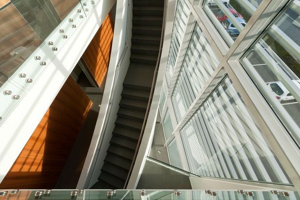 Upward-looking view of a contemporary multi-storey interior atrium with a central dark stairway, extensive floor-to-ceiling glazing, glass balustrades, white structural surfaces, and warm timber wall panelling. The space has strong vertical lines, abundant daylight, and a dramatic double-height character.