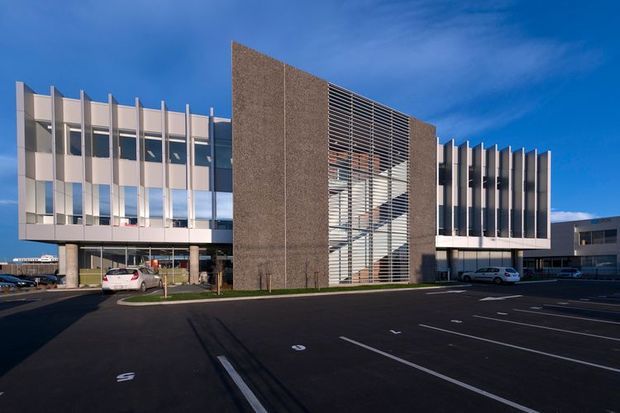 Exterior photograph of a contemporary multi-storey commercial or institutional building viewed from a large paved parking area. The facade combines extensive glazed curtain walls, projecting vertical fins, a tall textured dark masonry or concrete central volume, and a prominent glazed stair or atrium zone screened by horizontal louvers. The building has a crisp geometric composition and appears to be a large modern office, medical, or civic facility.
