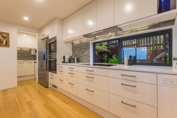Bright contemporary kitchen with extensive white built-in cabinetry, integrated stainless-steel appliances, a long benchtop, and a textured stacked-stone splashback. Warm timber flooring contrasts with the light joinery. A wide dark-framed window overlooks a planted courtyard, bringing greenery and natural light into the space.
