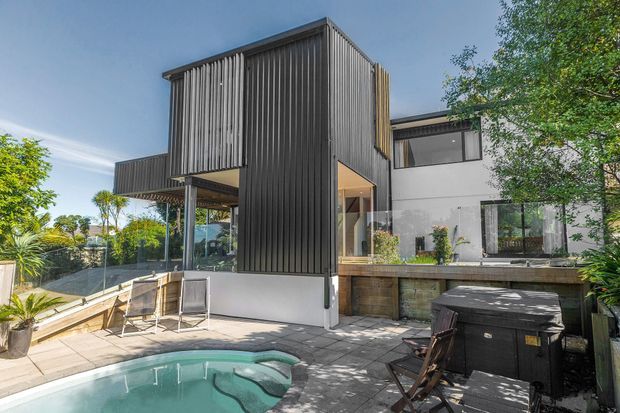 Exterior view of a contemporary multi-level residence with a strong vertical dark-clad addition, white rendered walls, large glazed openings, and elevated timber decking. The landscaped backyard includes a swimming pool, paved terrace, outdoor seating, and screening vegetation.