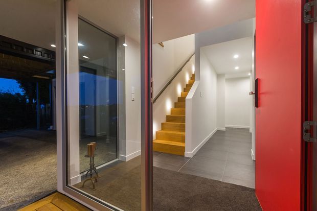 A modern residential entryway viewed from the open front door, with a vivid red door, full-height glazed side panel, dark tiled flooring, and a carpeted threshold. Inside, a straight timber staircase with a simple metal handrail leads upward against white walls, while recessed ceiling lights illuminate the hall. A small reflected decorative stand is visible in the glazing.