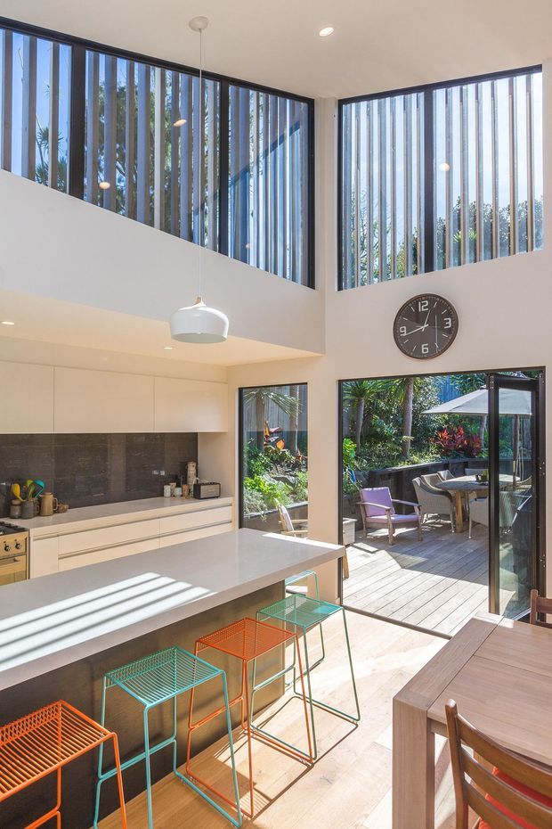 A bright, contemporary open-plan kitchen and dining area with a double-height corner glazed wall fitted with vertical blinds. The kitchen features white flat-panel cabinetry, a dark stone-look splashback, a pale island benchtop, and colourful wire bar stools. A dining table occupies the foreground, while large glazed doors open to a timber outdoor deck and leafy garden. A wall clock and pendant lighting add detail to the airy space.