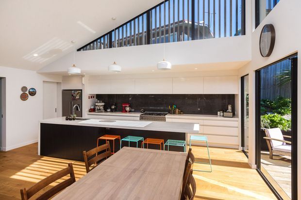 A bright, open-plan contemporary kitchen and dining area with a dramatic double-height void. The kitchen features extensive white flat-panel cabinetry, a large black island with a white benchtop, dark stone-look splashback, pendant lights, and bar stools. A timber dining table occupies the foreground, while floor-to-ceiling glazing and an upper-level balustrade bring in abundant natural light and overlook greenery outside.