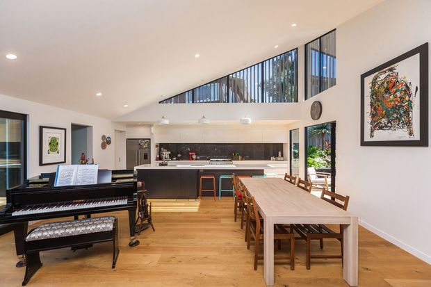 Bright, open-plan interior combining a kitchen, dining area and living/music zone. The space features warm timber flooring, a long dining table with wooden chairs, a black grand piano, dark kitchen cabinetry with a light island benchtop, and a high clerestory-style glazed opening overlooking greenery. Contemporary artwork and restrained furnishings add visual interest.