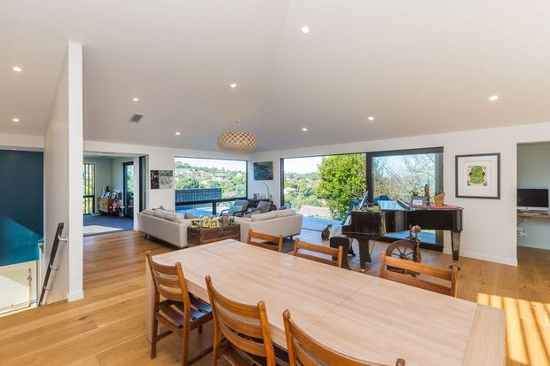 Bright open-plan living and dining area with polished timber flooring, a large timber dining table, mid-century style dining chairs, a white sectional sofa, grand piano, and expansive windows overlooking greenery. The room has a clean white ceiling with recessed lighting and a woven pendant over the dining zone, plus a glimpse of a stairwell and adjacent spaces.