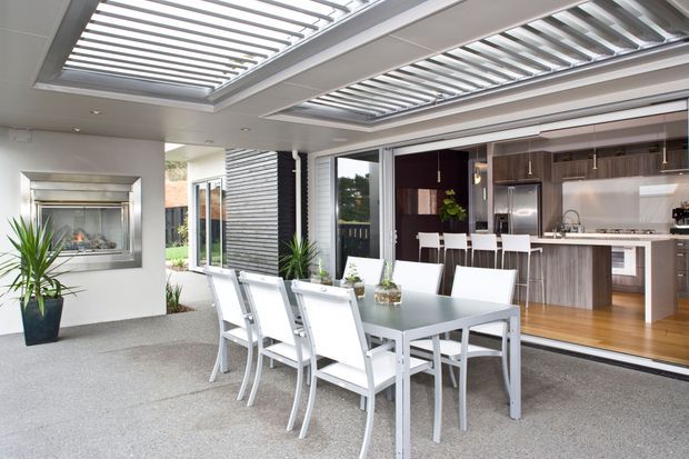 A contemporary open-plan outdoor entertaining area adjoining a modern kitchen. The covered patio features a large dining table with white chairs, a built-in wall fireplace, grey textured flooring, tropical potted plants, and extensive louvred roof panels that provide filtered daylight. The kitchen beyond has dark timber cabinetry, a pale benchtop, stainless-steel appliances, and timber flooring.