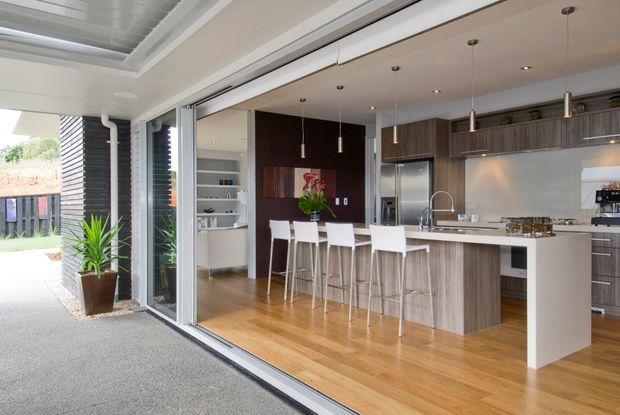Bright contemporary kitchen viewed from an adjoining outdoor or covered entertaining area through fully open sliding glass doors. The room features a long white island with waterfall end, integrated sink and seating for four, dark timber-grain cabinetry, stainless-steel appliances, pale grey splashback, pendant lighting, recessed ceiling lights, and warm timber flooring. An open-plan living or dining area is partially visible beyond the kitchen.
