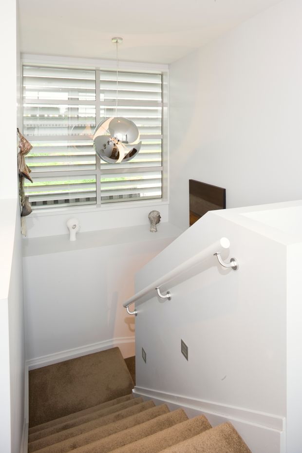Bright, minimalist stairway descending into a lower level, with warm beige carpeted steps, white walls and balustrade, brushed-metal handrail fittings, and a large horizontal louvred window providing natural light. A round pendant light and small decorative elements are visible near the landing.