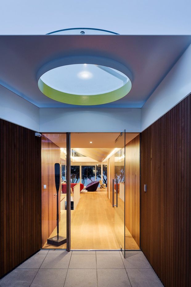 A contemporary entrance viewed from outside, featuring a covered porch with dark vertical timber wall cladding, large-format grey paving, and frameless glass entry doors. A sculptural circular ceiling light or skylight forms a prominent feature overhead. Through the doors is a warm timber-lined interior with polished timber flooring, decorative lighting, and colourful lounge seating.